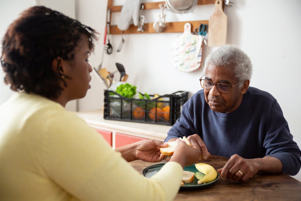 Home A woman assists an elderly man with meals at home, showcasing care and support.